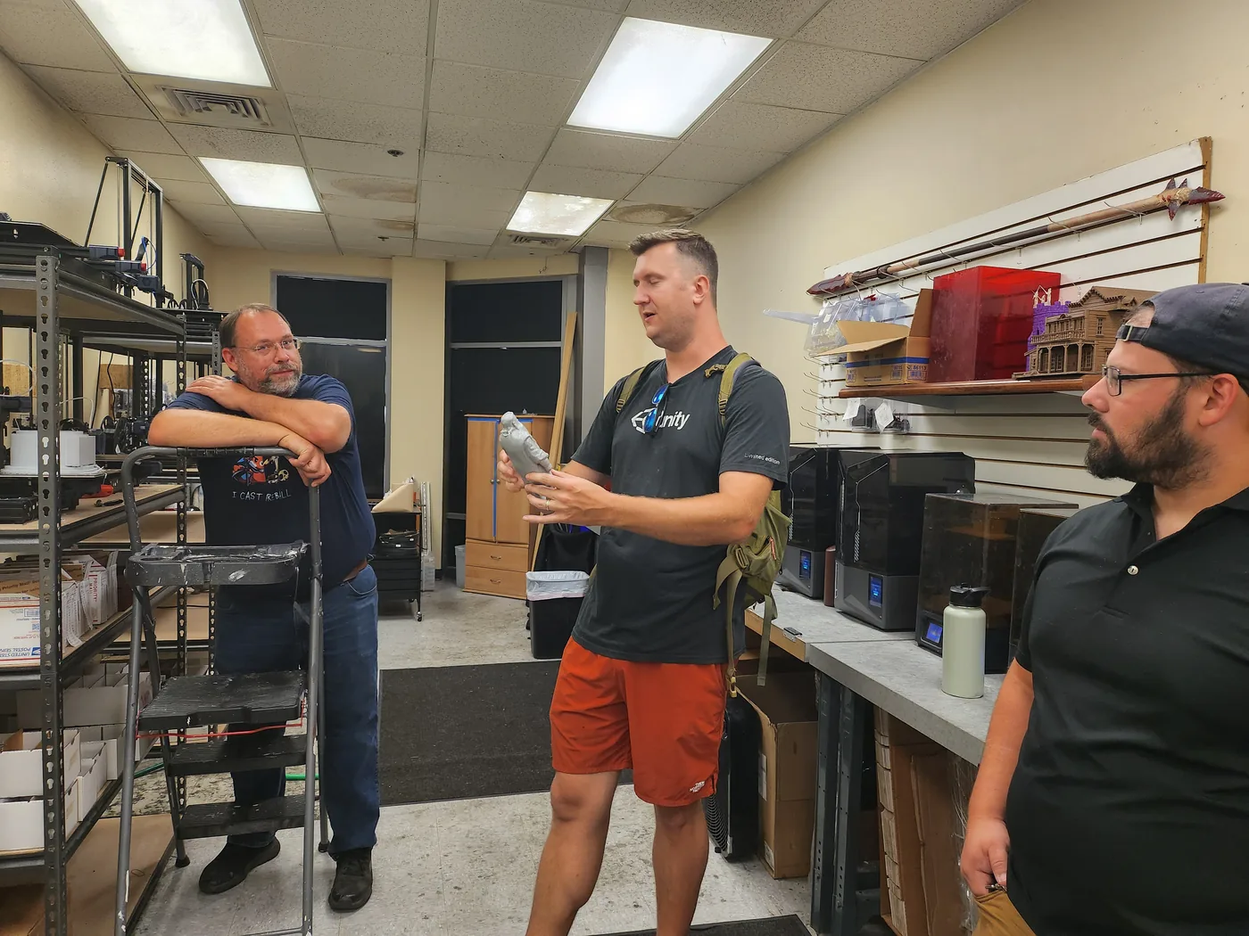 Three people talking in a room lined with 3D printers, one of them holding a freshly printed miniature
