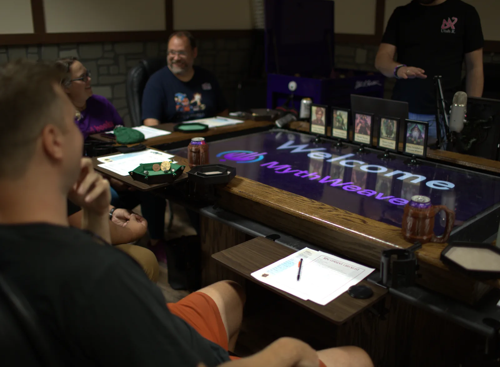 Four people around a large digital gaming table lit with the MythWeaver welcome screen, framed character portraits standing along the near edge